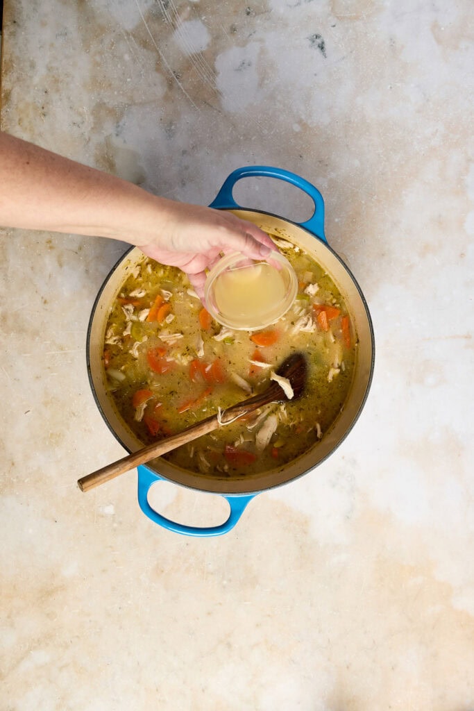 A hand pouring lemon juice into a pot of homemade lemon chicken rice soup, with vegetables and shredded chicken, on a rustic surface.