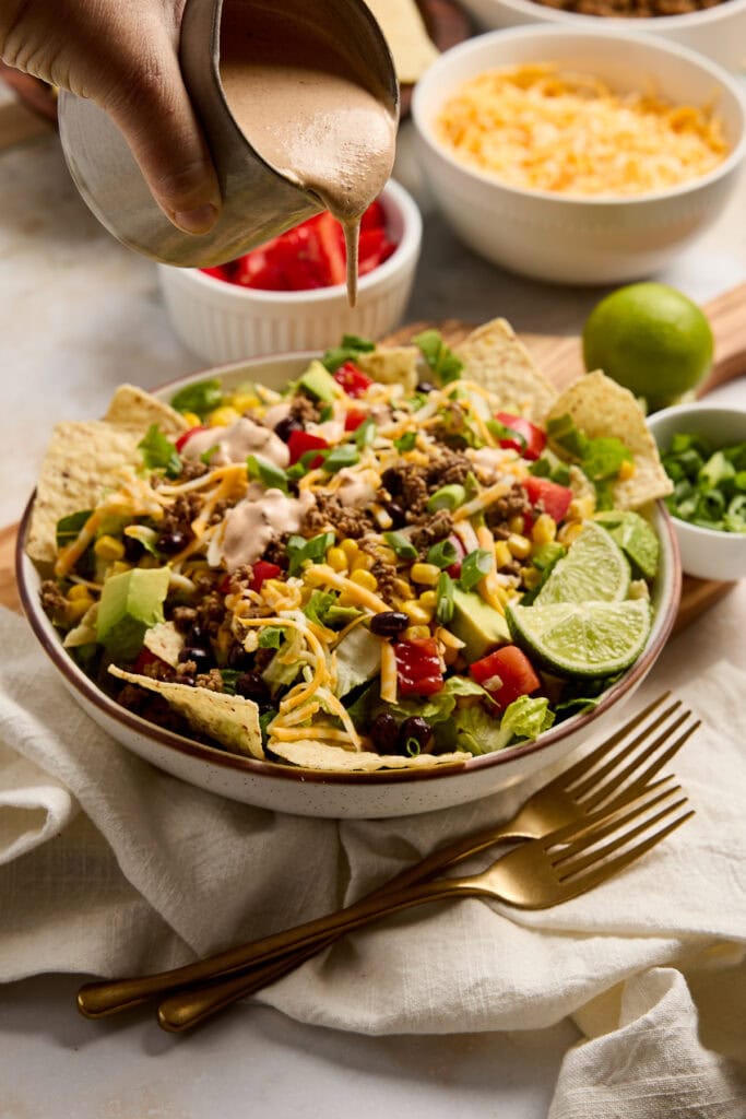 Colorful taco salad topped with dressing, cheese, and fresh lime slices, served in a white bowl with various toppings and ingredients around.