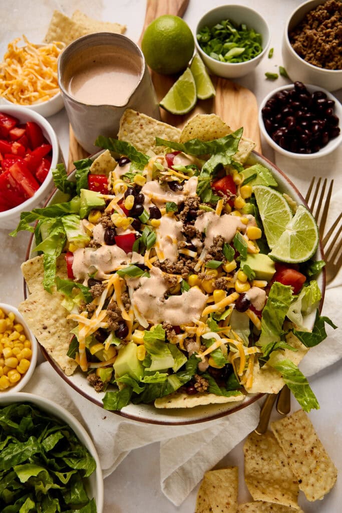 Colorful taco salad featuring ground beef, lettuce, corn, black beans, shredded cheese, and lime wedges, served with tortilla chips and various toppings.