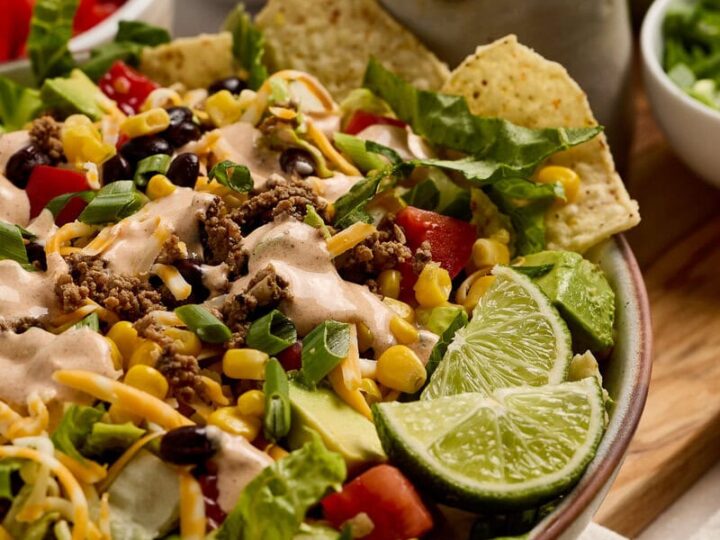 Colorful taco salad featuring fresh lettuce, tomatoes, shredded cheese, ground beef, corn, and crunchy tortilla chips, served with lime wedges and creamy dressing for a delicious meal.