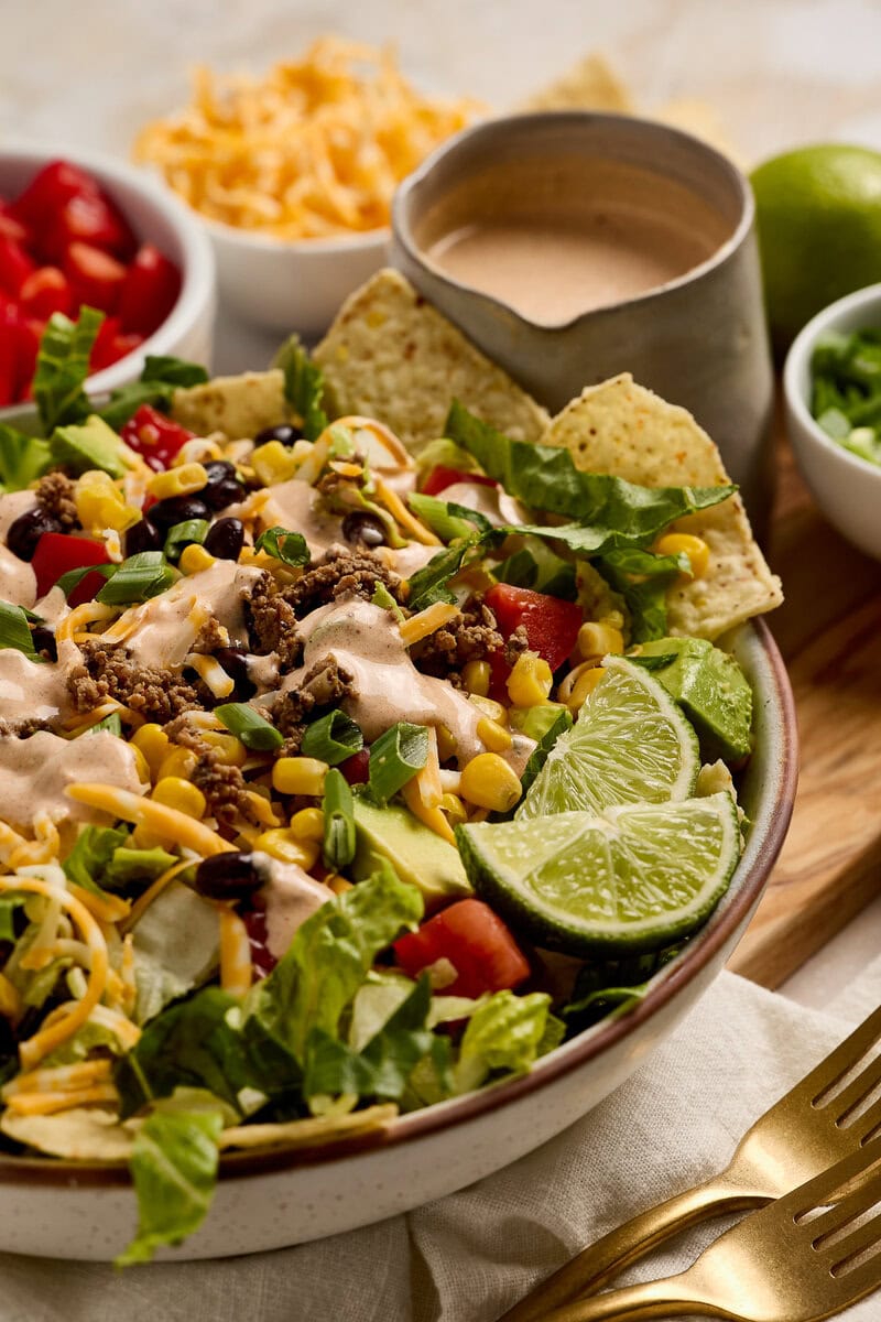 Colorful taco salad featuring fresh lettuce, tomatoes, shredded cheese, ground beef, corn, and crunchy tortilla chips, served with lime wedges and creamy dressing for a delicious meal.