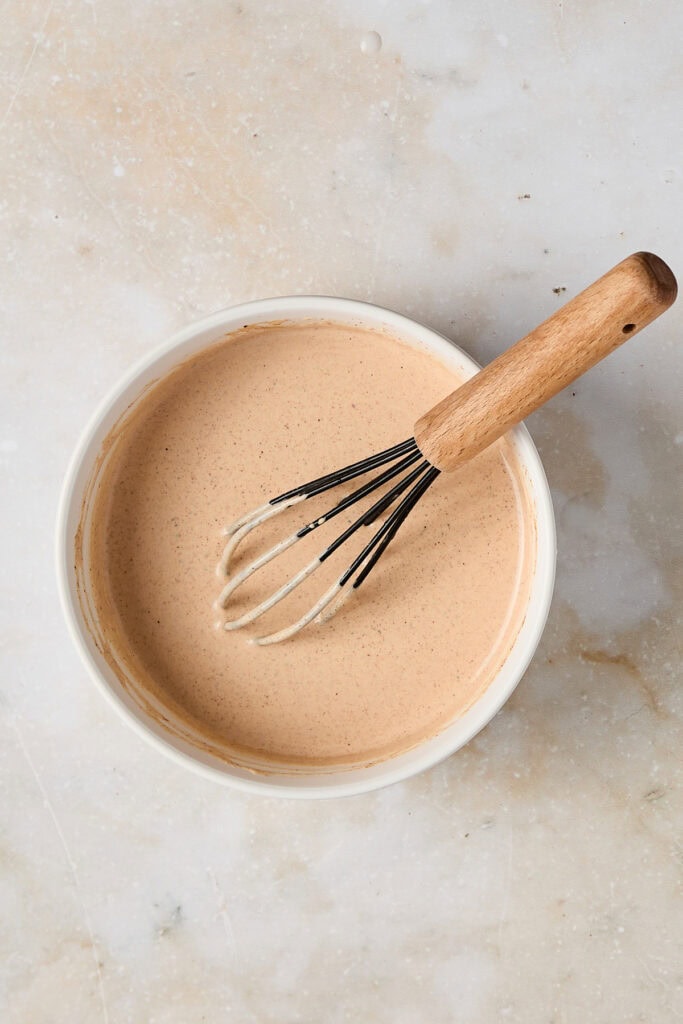 Creamy caramel sauce in a white bowl with a wooden-handled whisk, perfect for desserts and sweet recipes.