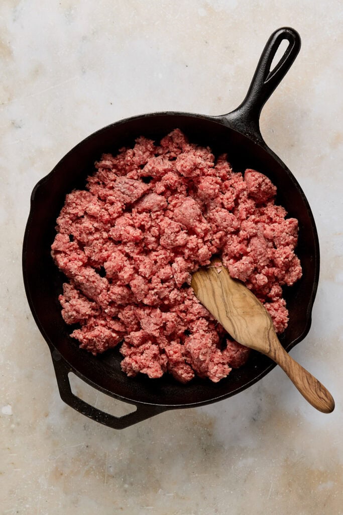 Ground beef cooking in a cast iron skillet with a wooden spoon.