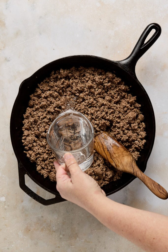 Delicious homemade chocolate cookie crumble being prepared in a skillet, with a hand pouring water and a wooden spoon ready for mixing. Perfect for dessert recipes or baking projects.