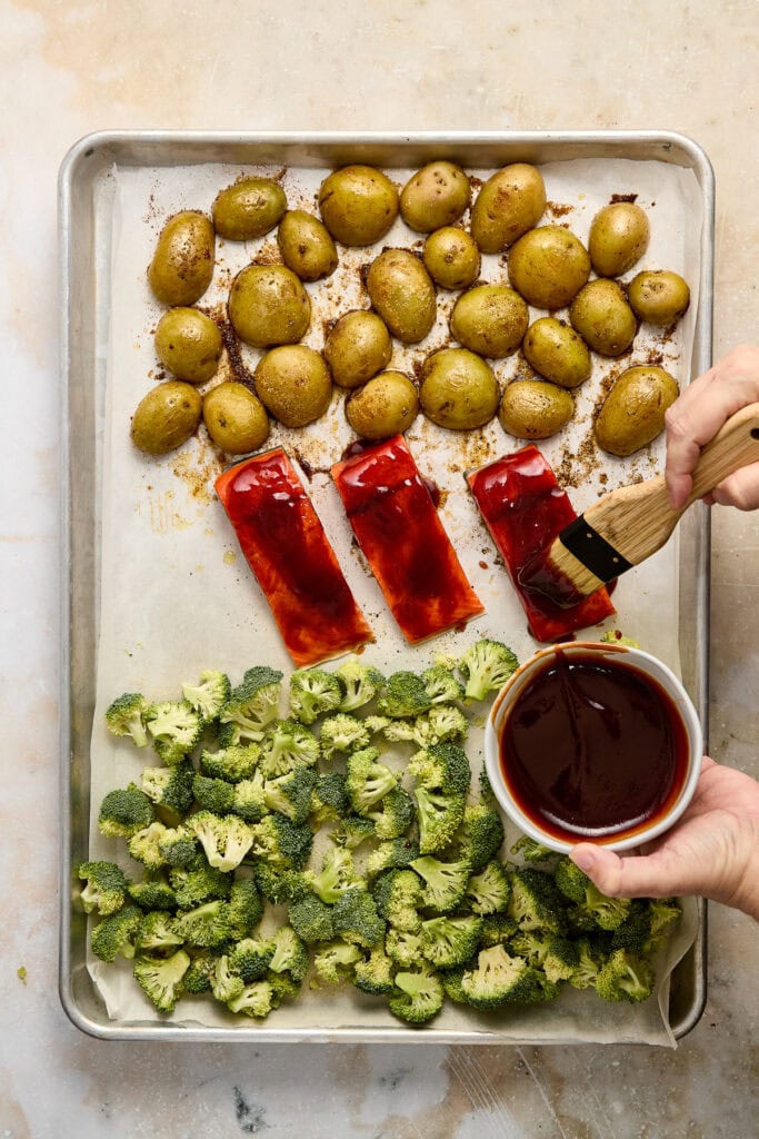 Fresh salmon slathered with barbecue sauce, potatoes, and broccoli on a baking sheet, ready for oven roasting. Perfect for nutritious meal prep, featuring fresh vegetables and vibrant colors.