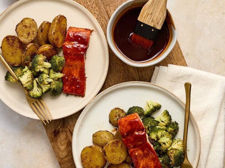 Barbecue salmon, roasted broccoli and potatoes on white plates, with a wooden cutting board and condiments in the background.