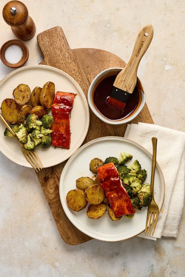 Barbecue salmon, roasted broccoli and potatoes on white plates, with a wooden cutting board and condiments in the background.