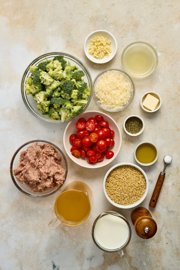 Assorted fresh ingredients for broccoli and cherry tomato salad, including broccoli florets, cherry tomatoes, grated cheese, and various dressings on a neutral background.