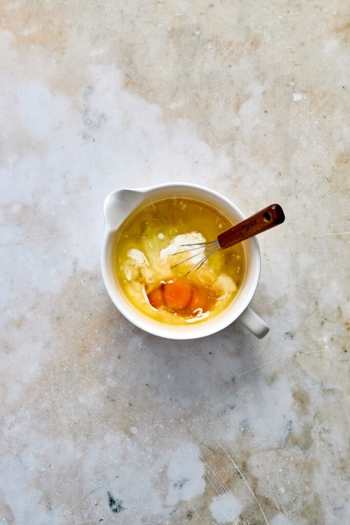 Egg custard with vanilla and caramel flavors in a white bowl, with a whisk resting inside, on a textured neutral background. Perfect for dessert or baking recipes.