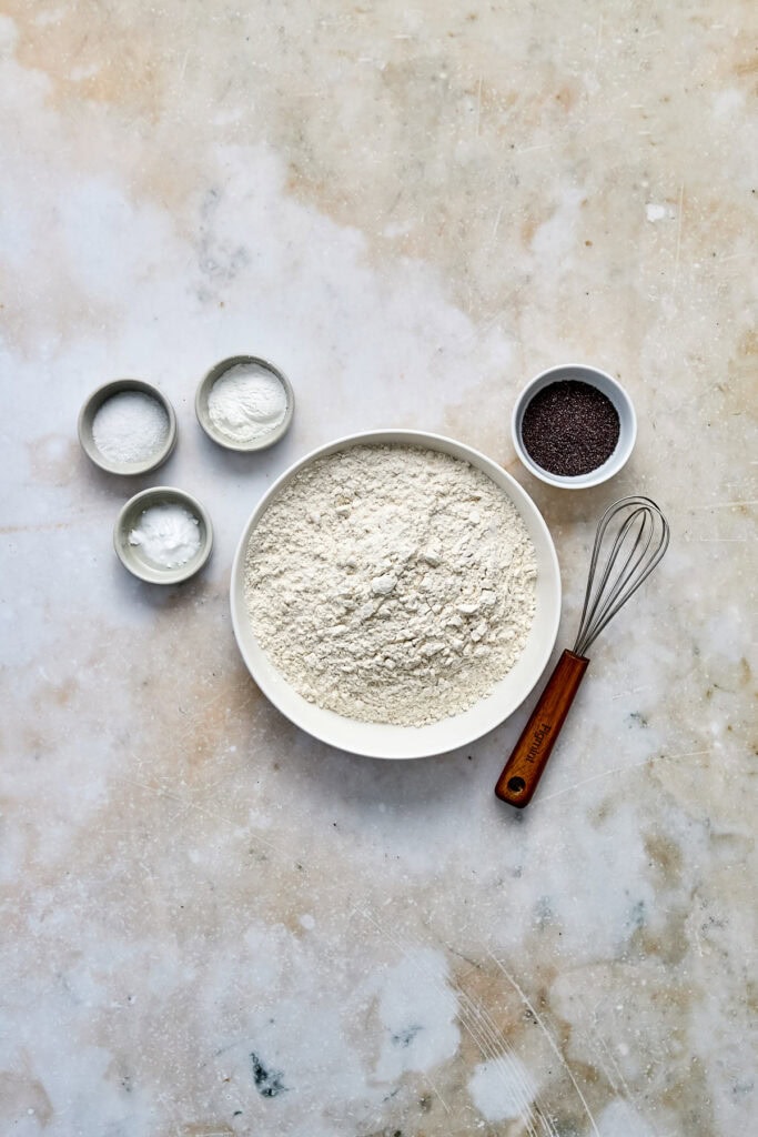 Assorted baking ingredients including flour, sugar, baking powder, and cocoa powder with a whisk on a textured surface. Perfect for baking and dessert recipes.