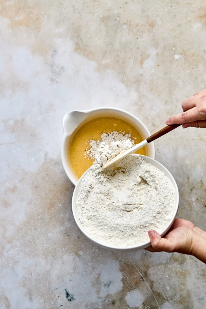 Close-up of hands mixing flour and wet ingredients in bowls for baking cookies, with a neutral textured background.