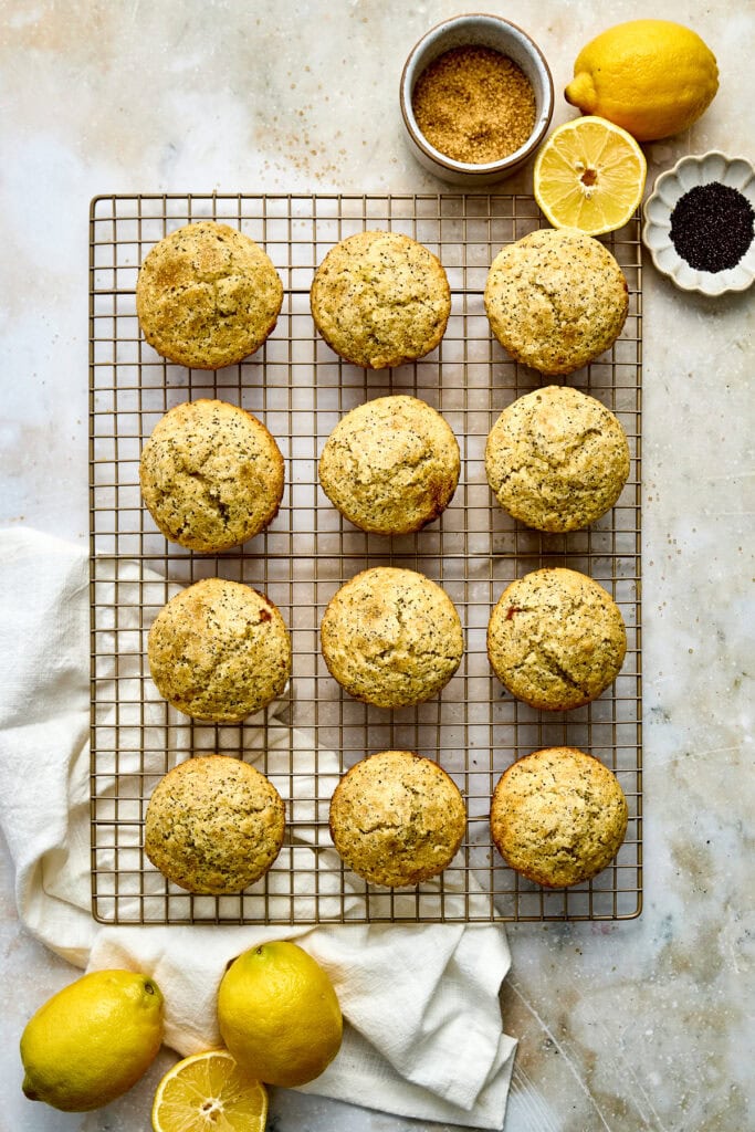 Delicious lemon poppy seed muffins cooling on a wire rack, with fresh lemons, lemon slices, and baking ingredients in the background, perfect for breakfast or snack.