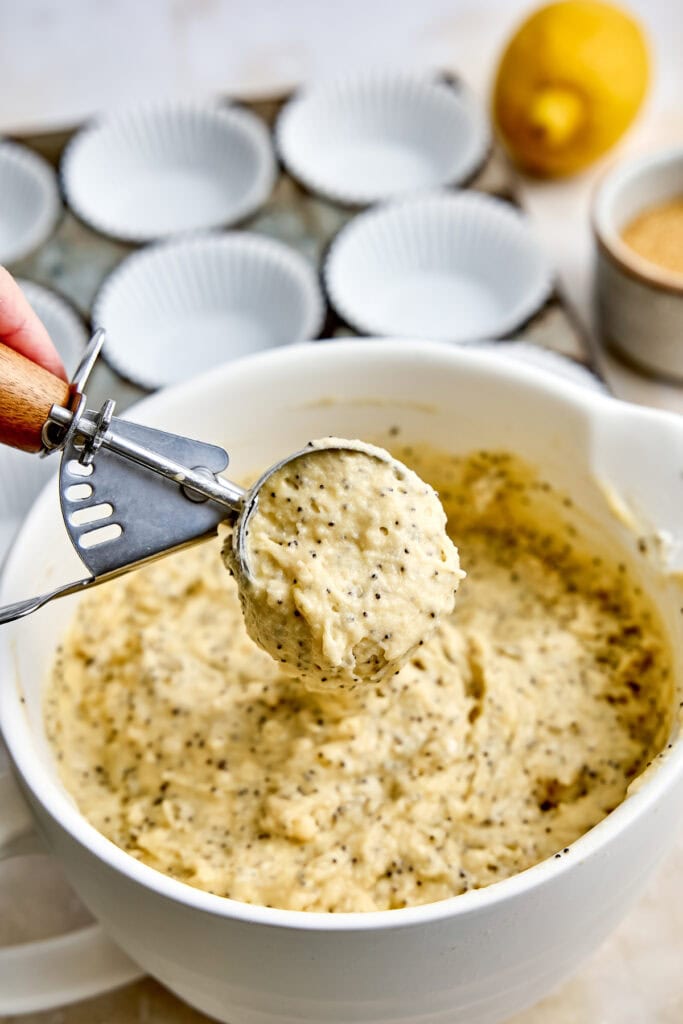 Close-up of lemon cake batter being scooped from a mixing bowl with a cookie scoop, surrounded by baking cups, lemon, and ingredients, ready for baking.