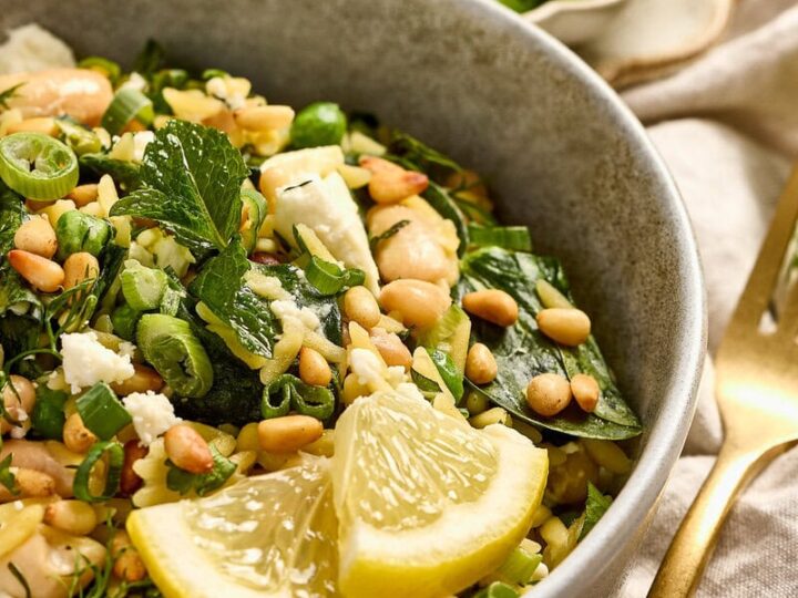 Close-up of a spinach feta orzo with lemon wedges and pine nuts in a gray bowl, garnished with herbs, on a neutral table setting.