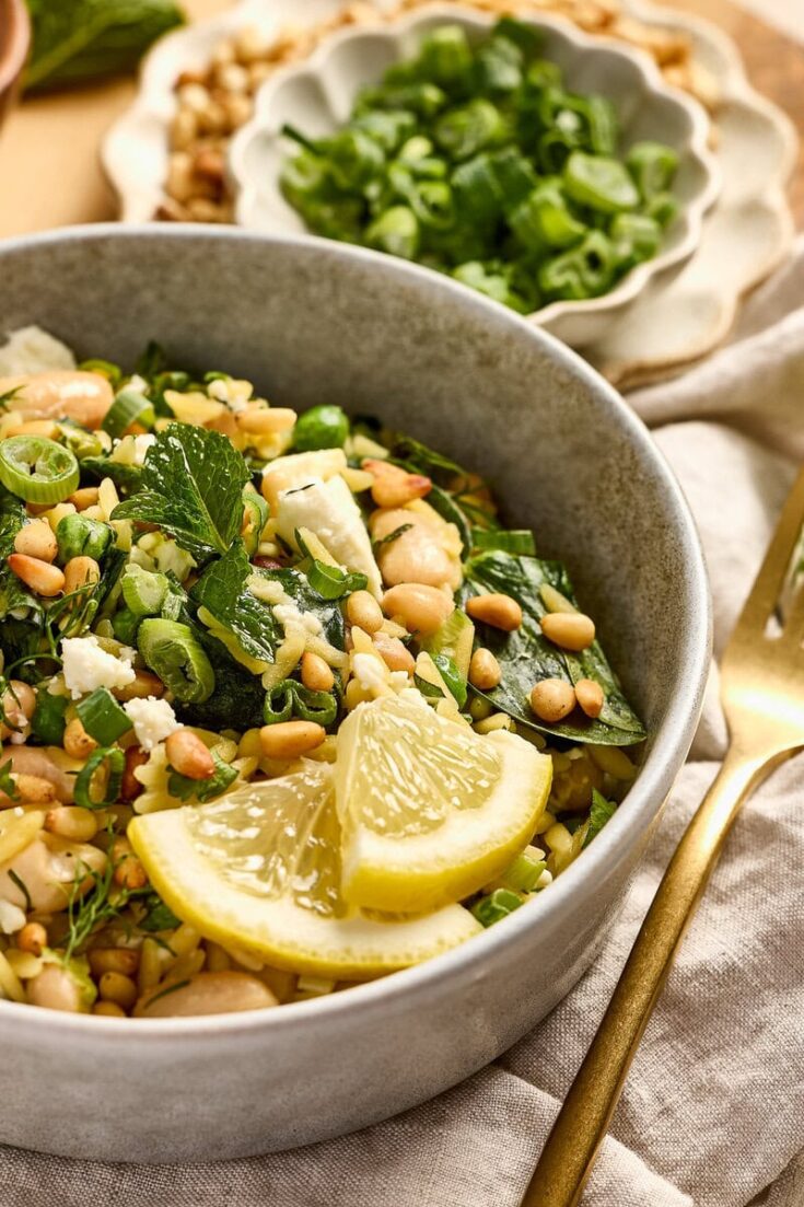 Close-up of a spinach feta orzo with lemon wedges and pine nuts in a gray bowl, garnished with herbs, on a neutral table setting.
