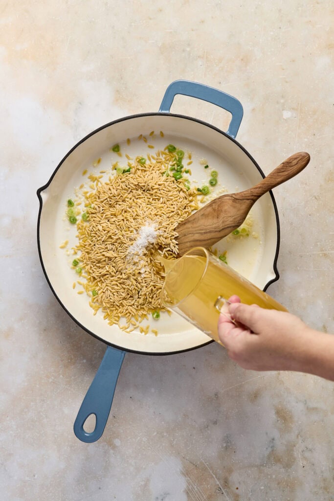 Delicious spinach feta orzo cooking in a creamy white sauce with white beans and fresh lemon zest, served in a blue skillet with a wooden spoon and a hand pouring broth or milk.