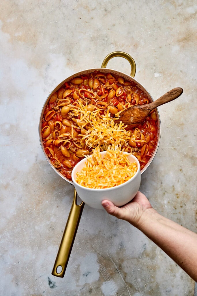 Hand holding a bowl of shredded cheese over a pot of taco pasta with tomato sauce, garnished with cheese, on a neutral background.
