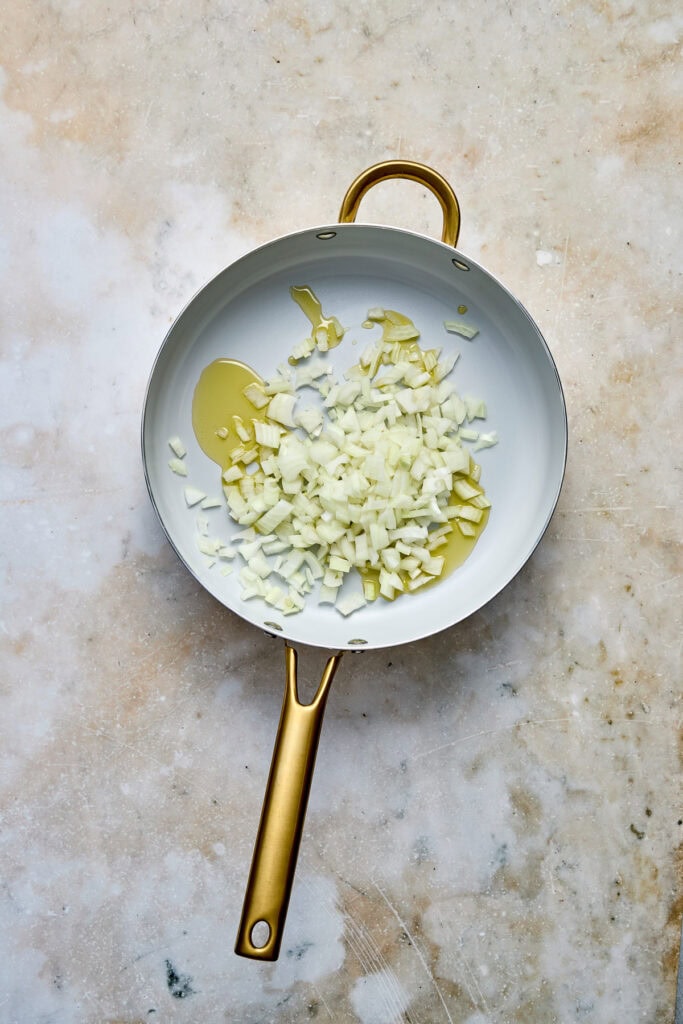 Olive oil and chopped onions in a white skillet with a gold handle, on a textured beige background.