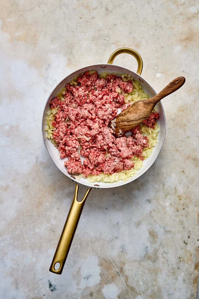 Close-up of a skillet with cooked ground beef and chopped onions, ready to serve, on a textured surface.