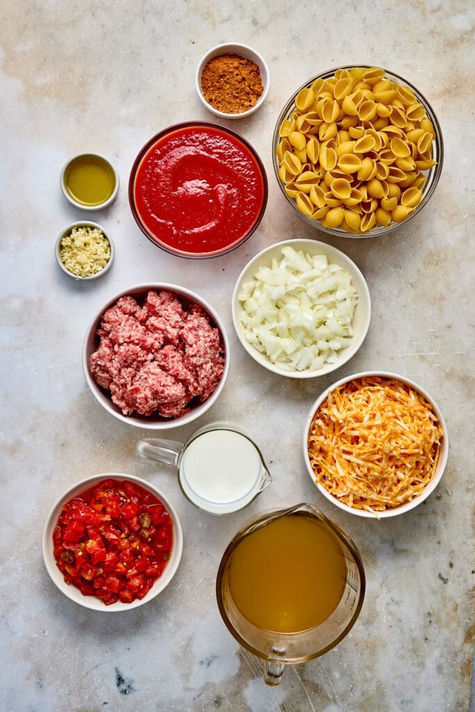 Assorted ground beef taco pasta ingredients including uncooked pasta, tomato sauce, ground meat, chopped onions, diced tomatoes, cheese, and seasonings arranged on a light surface.