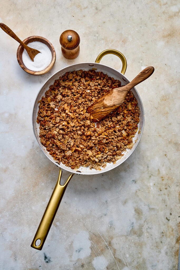 Close-up of cooked ground beef mixture in a skillet with a wooden spoon, salt, and pepper shakers on a neutral background.