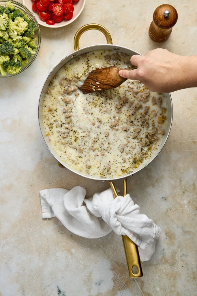 Creamy cheese sauce cooking in a skillet with fresh broccoli and cherry tomatoes nearby, perfect for a delicious meal or snack.