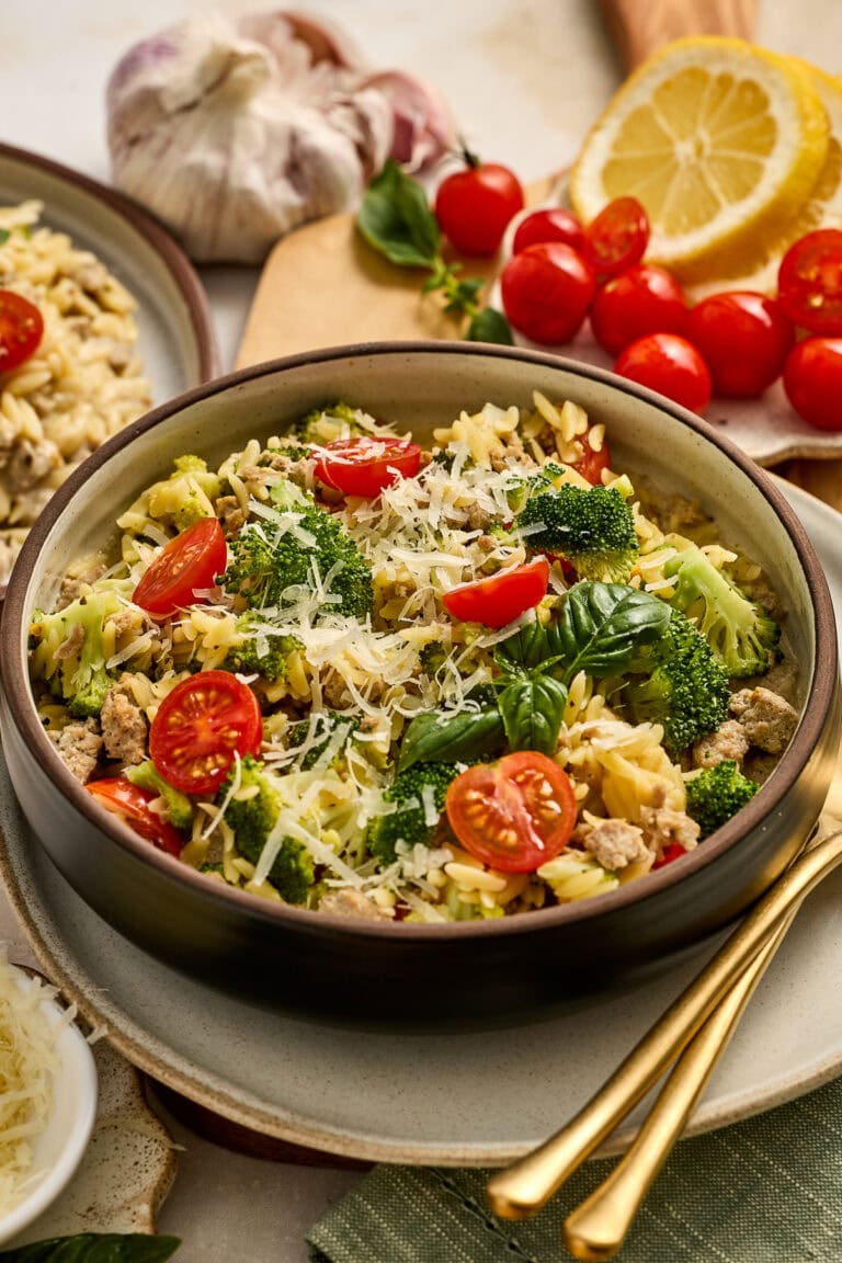 Fresh vegetable pasta salad with cherry tomatoes, broccoli, and grated Parmesan cheese, served in a bowl with lemon slices and garlic in the background. Perfect for healthy, flavorful meals.