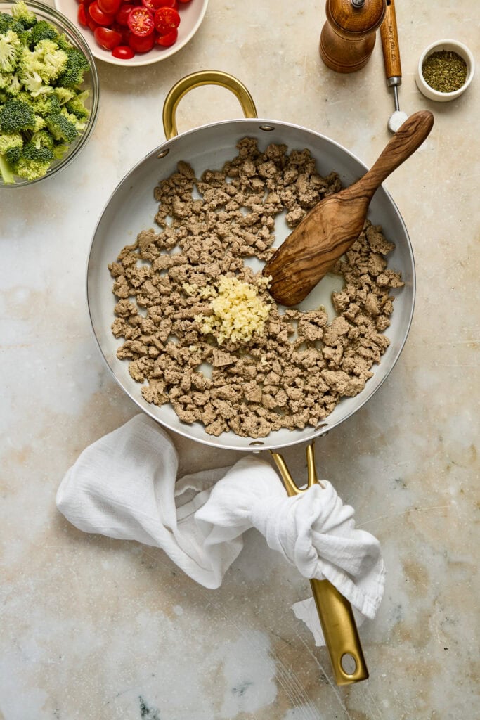 Ground beef cooked with garlic in a skillet, ready for a delicious meal, with fresh vegetables and seasonings nearby.