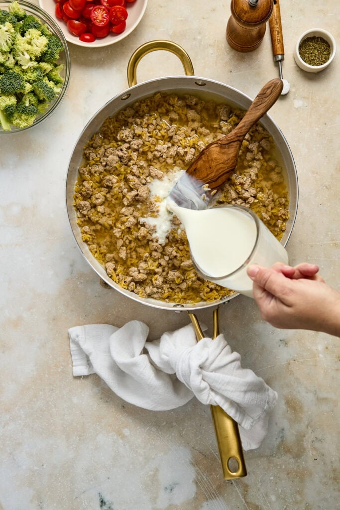 Close-up of a skillet with cooked sausage and rice, being poured with cream, surrounded by fresh broccoli, cherry tomatoes, and seasonings on a light kitchen countertop.