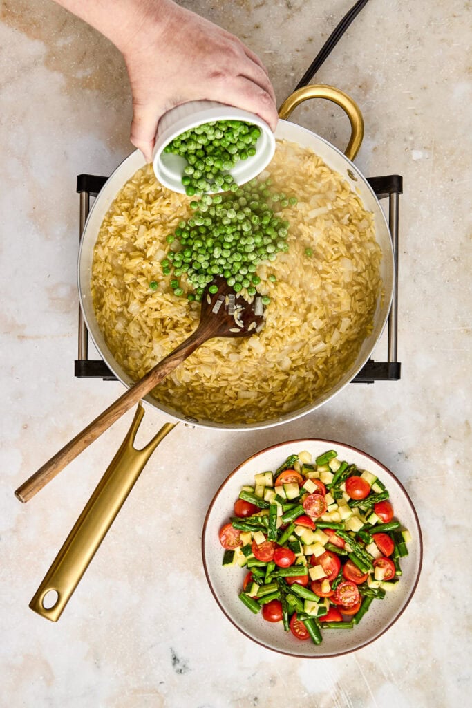A hand adding green peas to a creamy risotto in a skillet, with a bowl of fresh vegetable salad nearby on a neutral countertop.