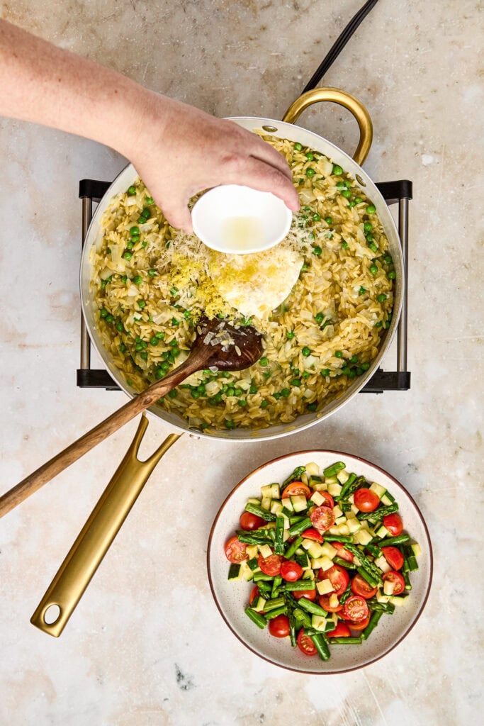 A hand pouring cream into a pan of freshly cooked risotto garnished with herbs, with a side bowl of chopped tomatoes, cucumbers, and green beans on a neutral surface.