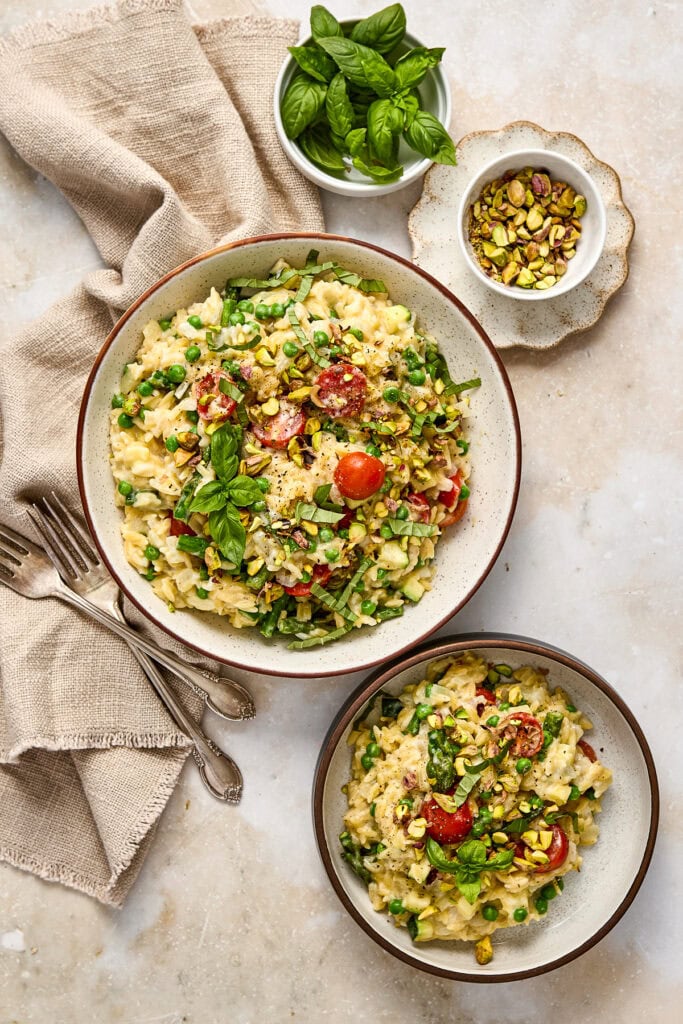Delicious creamy orzo primavera garnished with fresh basil, cherry tomatoes, and chopped pistachios, served in rustic bowls for a flavorful summer side dish.