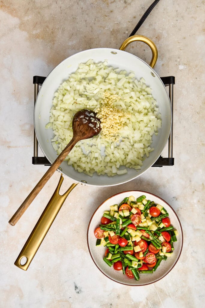 Sautéed onions and garlic in a white skillet with a wooden spoon, paired with a colorful vegetable salad of cherry tomatoes, cucumbers, and green beans in a bowl, on a neutral surface.