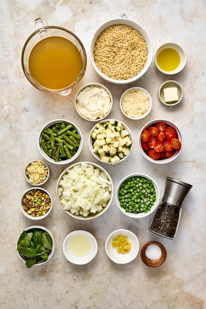 Assorted fresh ingredients for creamy pasta salad including pasta, cherry tomatoes, cucumbers, green peas, onions, herbs, and dressings arranged on a light surface.