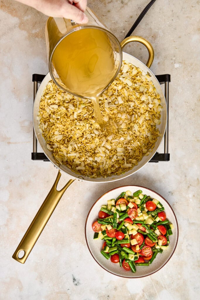 Pouring golden broth over a skillet of cooked chicken and vegetables, with a side bowl of fresh cherry tomatoes and chopped cucumbers, on a neutral countertop.