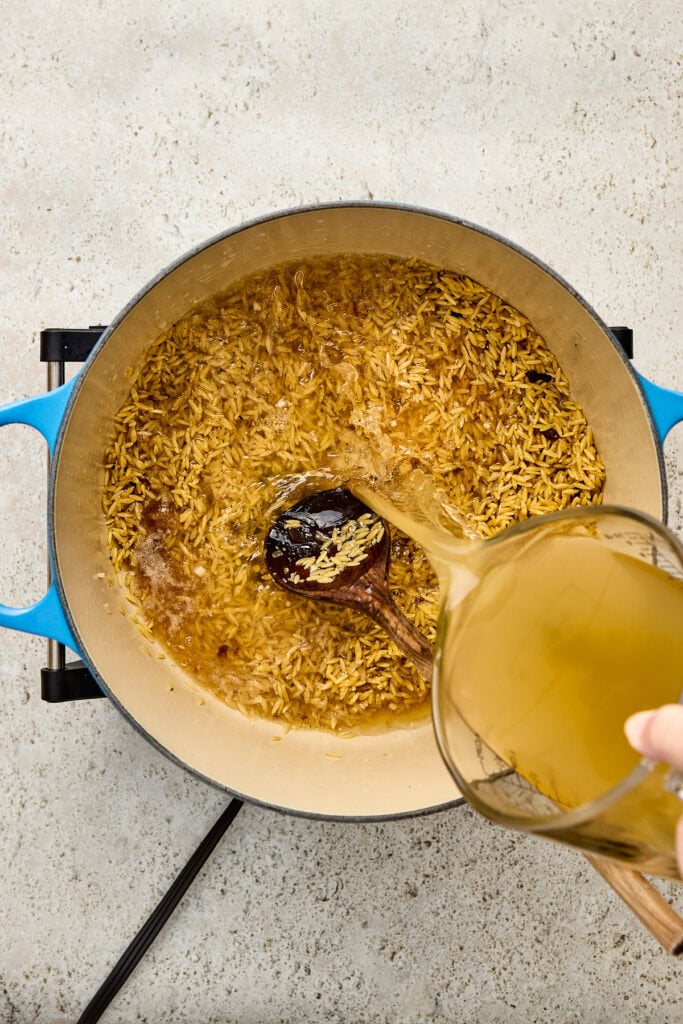 Cooking creamy chicken and rice in a blue skillet with broth being poured in, showcasing a delicious and hearty meal preparation.
