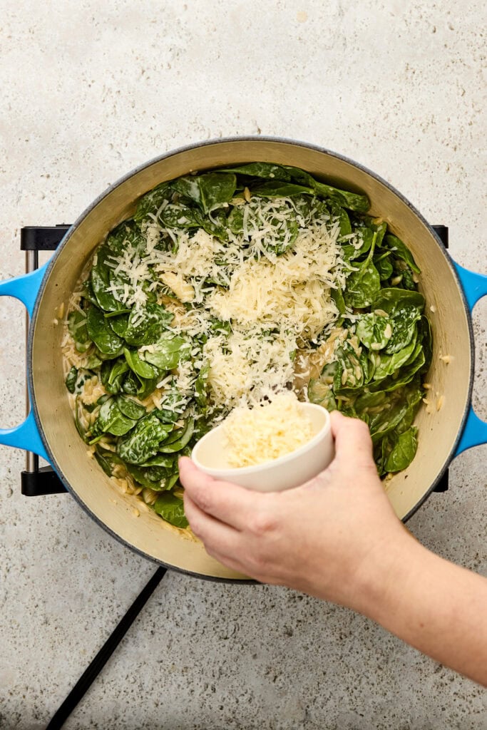 Cooking creamy spinach and cheese pasta in a blue skillet, with grated cheese being added from a small bowl, on a light countertop.