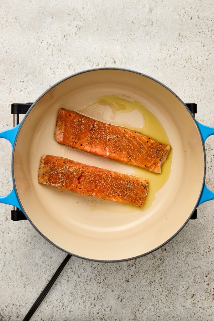 Two seasoned salmon fillets cooking in a blue skillet with olive oil, on a light textured surface, showcasing fresh seafood preparation.