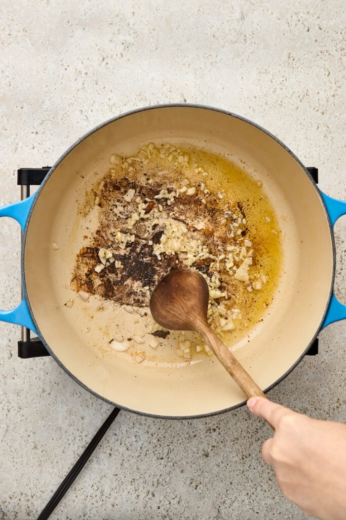 Close-up of garlic butter sauce simmering in a blue pot, with a wooden spoon stirring the mixture, on a light textured surface.