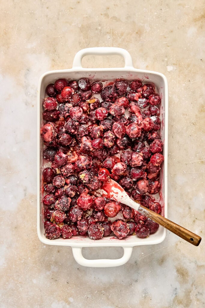Frozen mixed berries in a white baking dish with a wooden spoon, ready for baking or cooking, on a neutral surface.