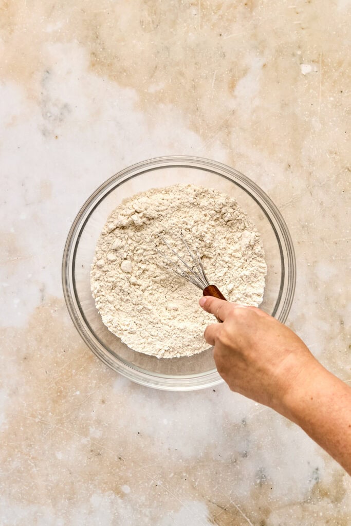 Close-up of a hand whisking flour in a glass bowl on a textured surface, preparing for baking at Stuck On Sweet.