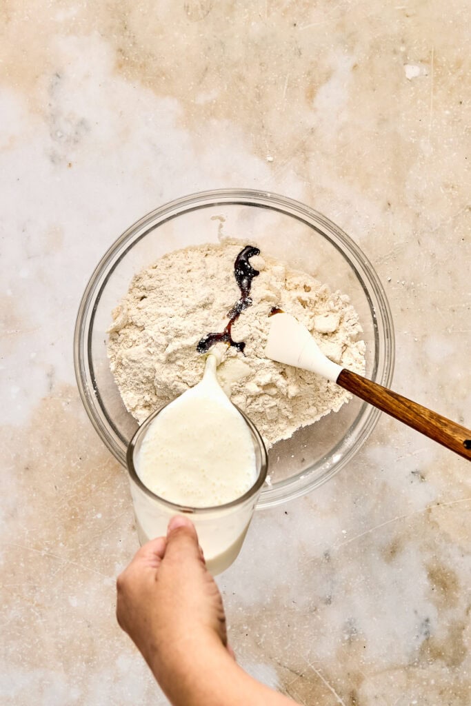 Pouring milk into a bowl of flour and cocoa powder for baking, with a wooden spoon on a textured surface.