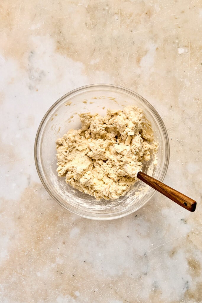 Close-up of cookie dough in a glass bowl with a wooden spoon on a light textured surface, perfect for baking and dessert recipes.