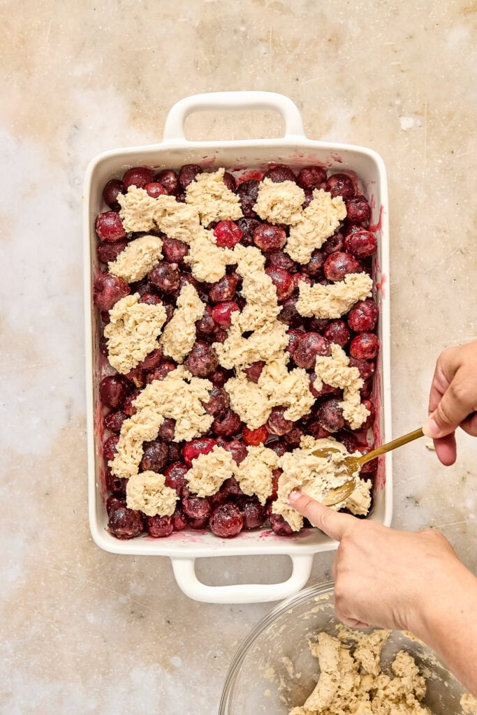 A person preparing a mixed berry crisp in a white baking dish, with fresh berries and crumbly topping, ready to be baked or served.