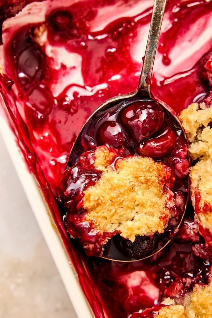 Close-up of a cherry cobbler dessert with a spoonful showing cherries, berries, and a crumbly topping, perfect for sweet cravings.