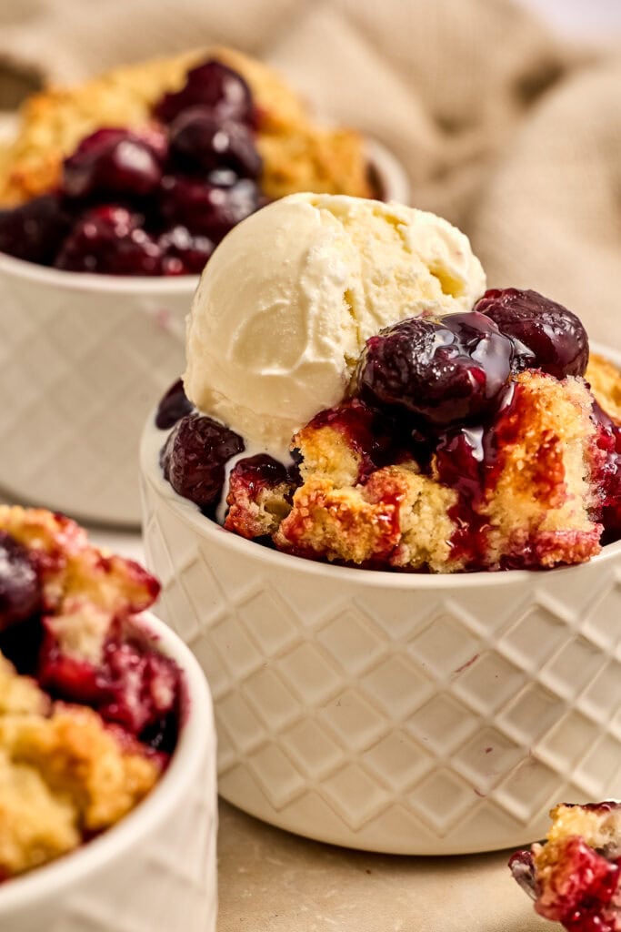 Delicious cherry cobbler topped with a scoop of vanilla ice cream in a white bowl, served with fresh cherries and warm, crumbly crust.