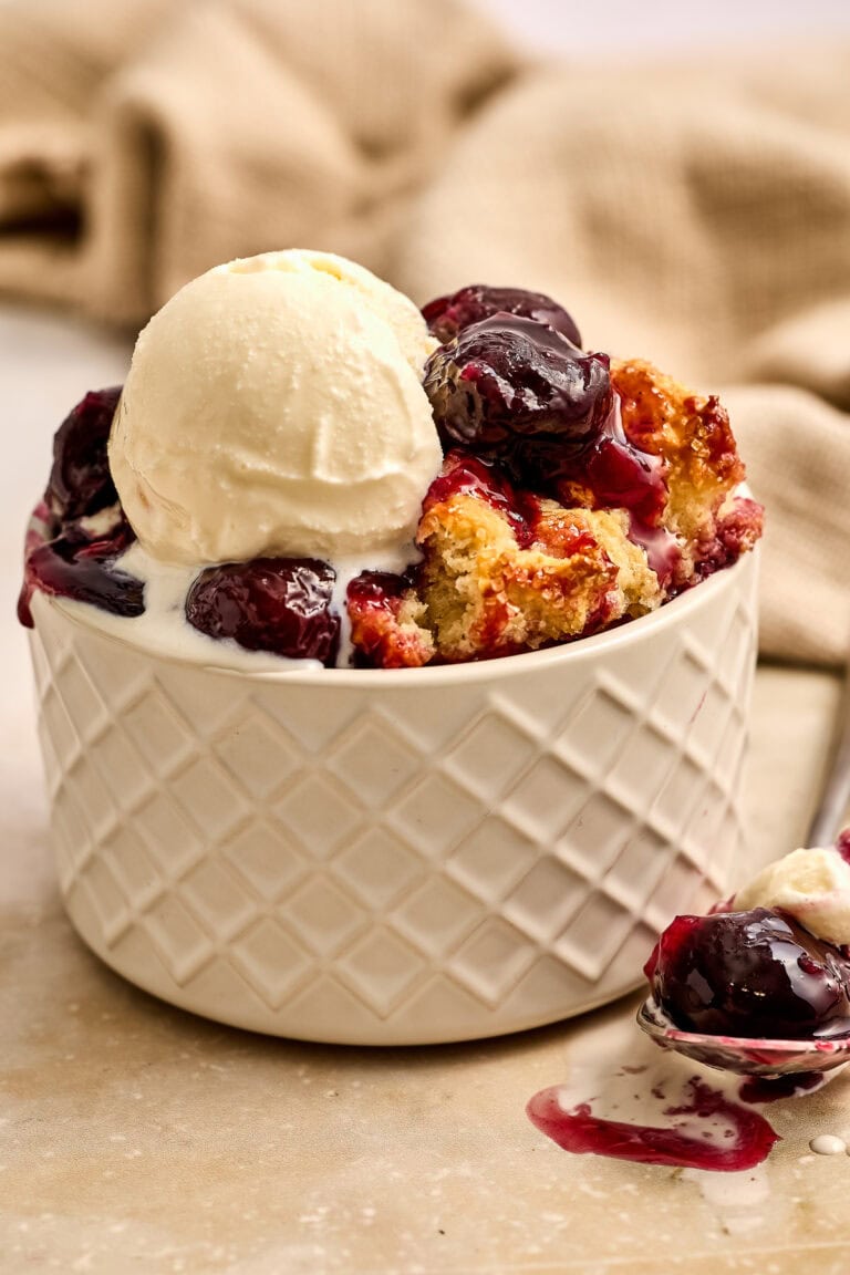 Delicious cherry cobbler topped with a scoop of vanilla ice cream in a white bowl, garnished with fresh cherries and warm berry syrup.