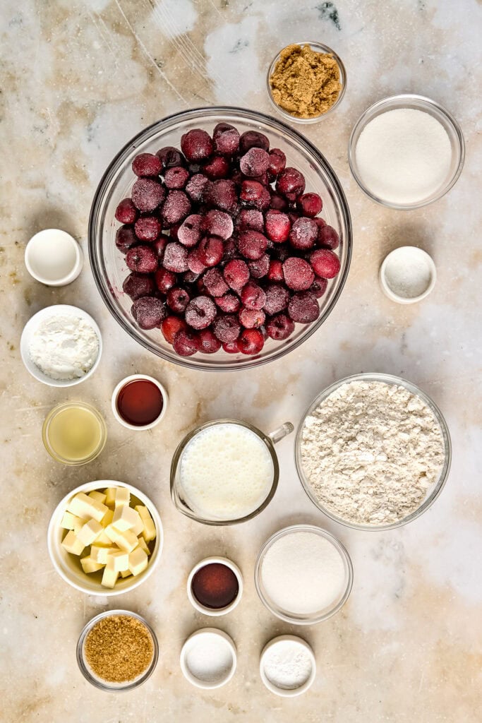 Assorted ingredients for making a cranberry pie, including fresh cranberries, sugar, flour, butter, and spices, arranged on a marble surface.