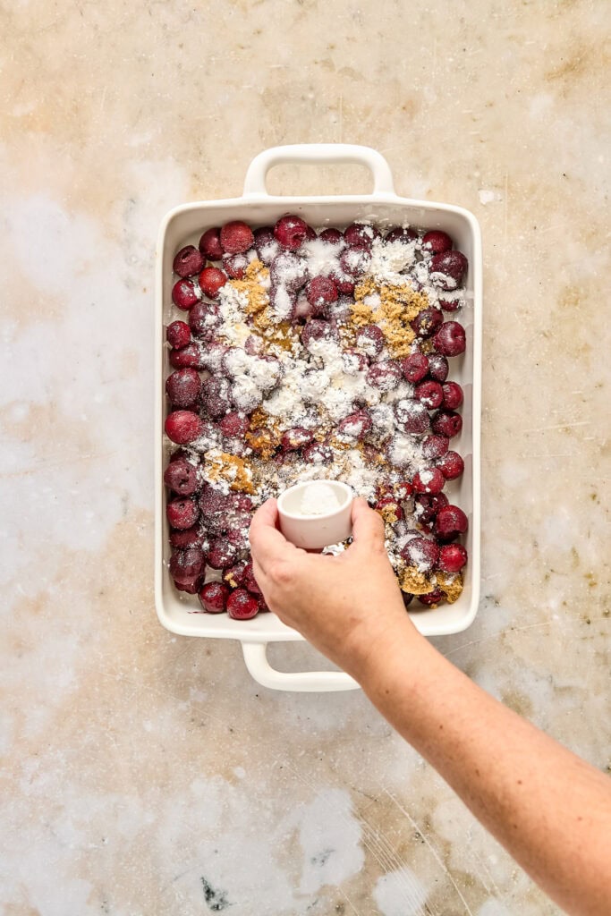 Hand sprinkling powdered sugar and crumble topping over fresh berries in a baking dish for a dessert.