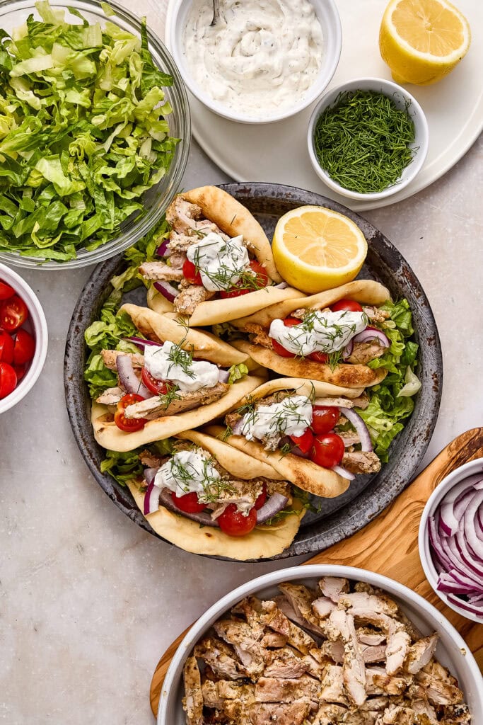Delicious Mediterranean-style greek chicken pitas topped with cherry tomatoes, red onions, and fresh herbs, served with lemon wedges and creamy sauce on a gray surface.